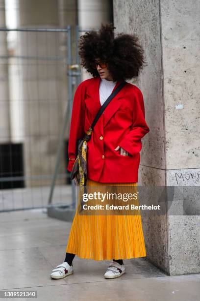 Guest wears sunglasses, a white t-shirt, a red oversized blazer jacket, an orange pleated / accordion midi skirt, black tights, a black shiny leather...
