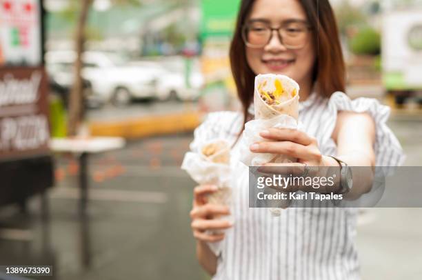 a southeast asian woman is holding take-out wrap sandwich outdoors - shawarma stock-fotos und bilder