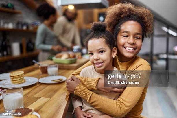 glückliche afroamerikanische schwestern, die sich im esszimmer umarmen. - sitting at table looking at camera stock-fotos und bilder