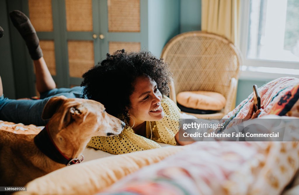 Beautiful woman lies on her stomach on a colourful bed, beside her lurcher dog, and takes a selfie.