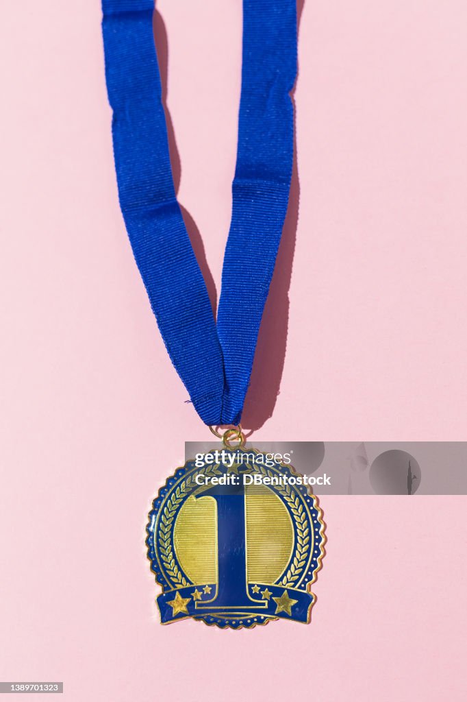 First place champion medal with number 1 in blue and blue strap on a pink background. Concept of competition, winner, winner, sport, triumph, champion and first place.