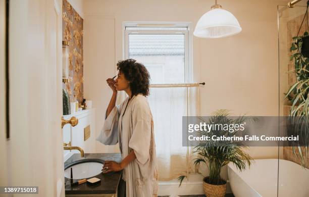 beautiful black woman applies make-up in a bathroom mirror - applying imagens e fotografias de stock