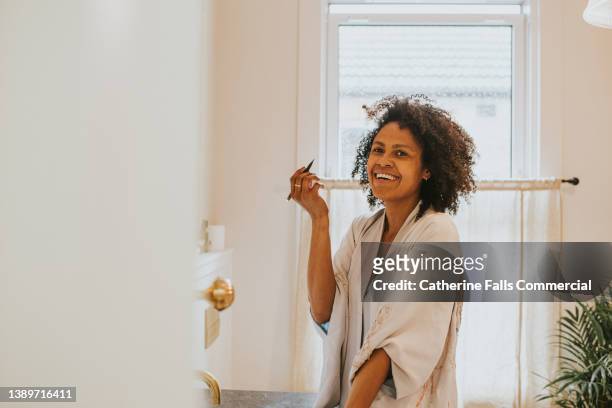 beautiful black woman applies make-up at a bathroom mirror. she looks over towards the camera and giggles - パウダールーム　女性 ストックフォ�トと画像