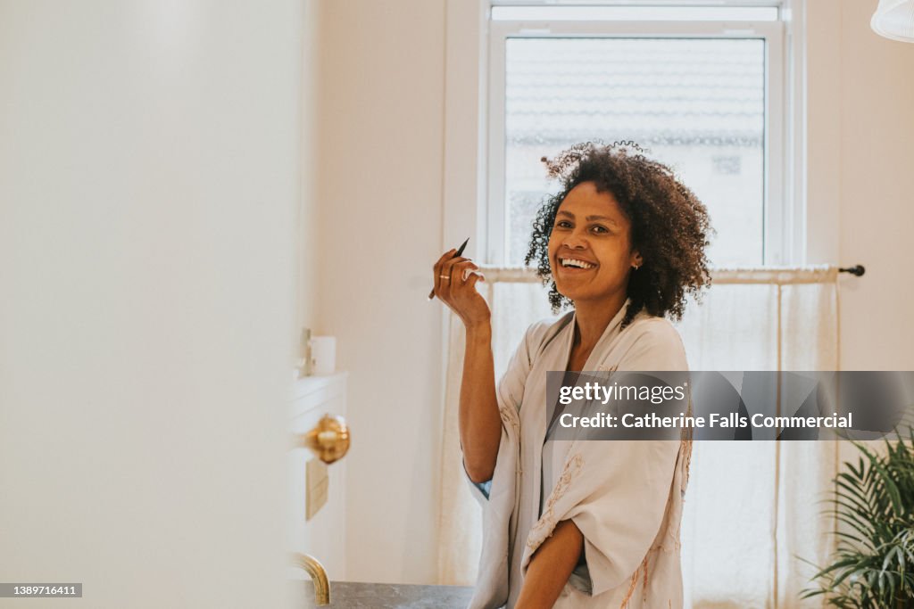 Beautiful black woman applies make-up at a bathroom mirror. She looks over towards the camera and giggles
