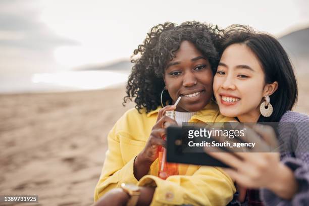 two friends sitting on the beach drinking soda and taking a selfie - asian female friends drinking soda outdoor stockfoto's en -beelden