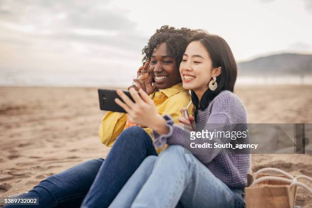 two friends sitting on the beach drinking soda and taking a selfie - asian female friends drinking soda outdoor stockfoto's en -beelden