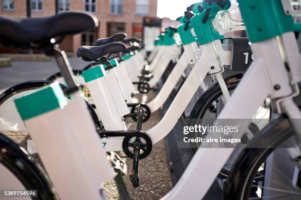 modern electric rental bikes lined up during charging on a sunny day, identified by number, brick building in background. - e bike stock pictures, royalty-free photos & images