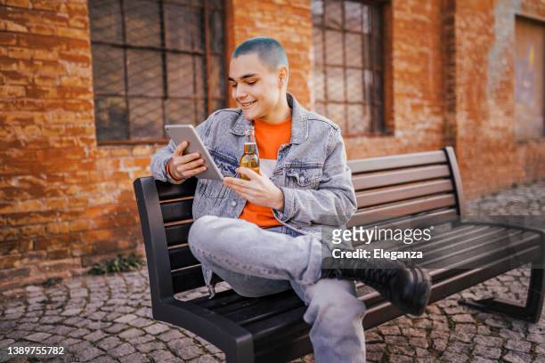 hipster junger mann, der sein digitales tablet benutzt und bier auf der parkbank trinkt - tablet alcohol stock-fotos und bilder