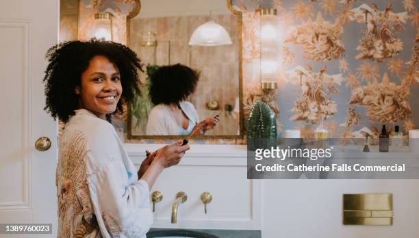 a beautiful woman glances over her shoulder as she applies make-up in a bathroom - standing mirror stock pictures, royalty-free photos & images