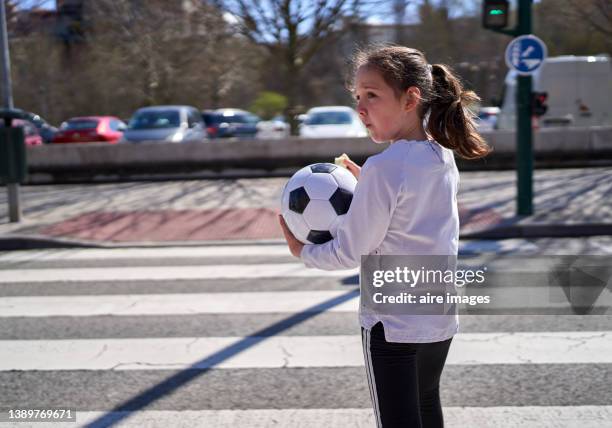 girl holding soccer ball while eating a snack, standing on the street in front of the crosswalk at midday. - 8 ball stock-fotos und bilder