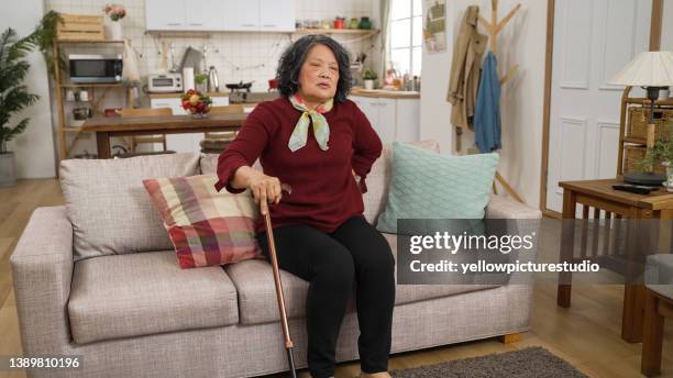 asian retired old woman struggling to stand up with a stick after fall on living room floor at home. she leans on a pillow while sitting and resting on the couch - fast forward stock pictures, royalty-free photos & images