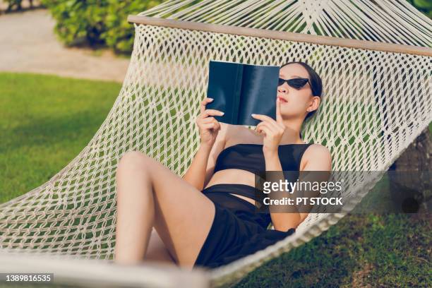 a woman wearing a black bikini and wearing a long black skirt, wearing black sunglasses, lies reading in a hammock. - hammock asia stock pictures, royalty-free photos & images