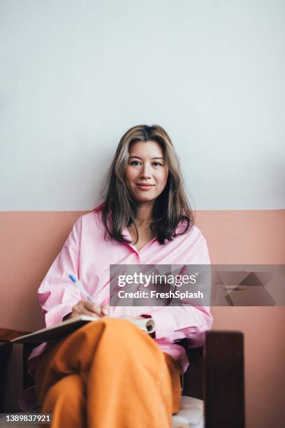 beautiful thai woman wearing a pink shirt and orange trousers sitting in her living room and writing her diary - pink collared shirt stock pictures, royalty-free photos & images