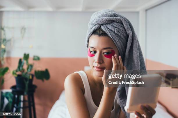 beautiful thai woman looking at herself in a hand mirror while applying under-eye patches - eye patch stock pictures, royalty-free photos & images