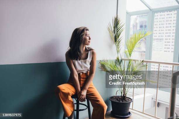 hermosa mujer tailandesa sentada en una silla de bar y mirando por la ventana - pantalón mujer fotografías e imágenes de stock