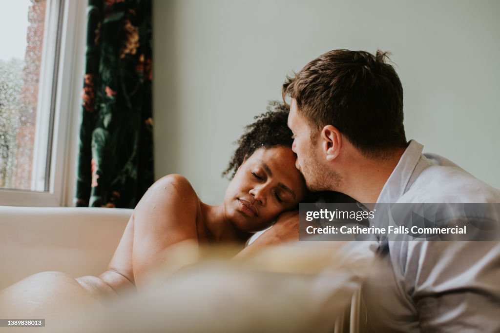 Loving image of a couple in a bathroom. The woman lies in a bathtub and places her head gently on her partners shoulder as he kisses her forehead.