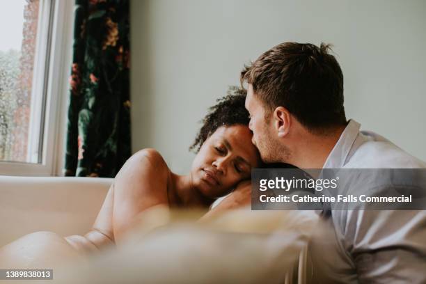 loving image of a couple in a bathroom. the woman lies in a bathtub and places her head gently on her partners shoulder as he kisses her forehead. - prop stock pictures, royalty-free photos & images
