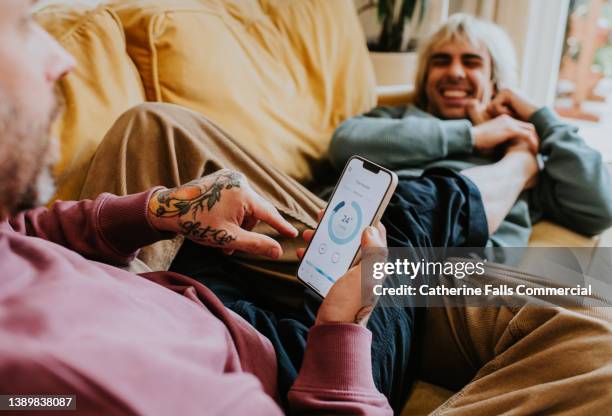 a couple reclining on a sofa use a phone app to control their thermostat - couple couch imagens e fotografias de stock