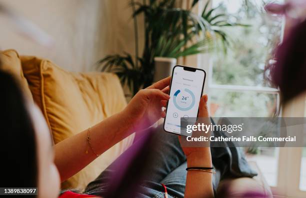 relaxed woman reclines on a sofa and uses a thermostat app on her smart phone to control the housing heating system - fuel efficient photos et images de collection