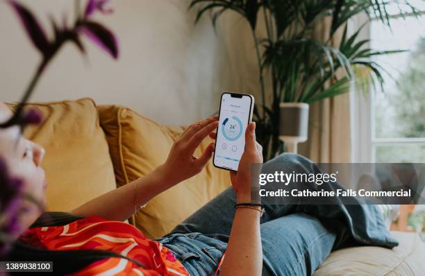 relaxed woman reclines on a sofa and uses a thermostat app on her smart phone to control the housing heating system - application smartphone photos et images de collection
