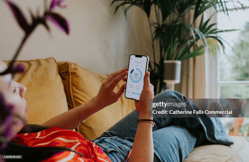 Relaxed woman reclines on a sofa and uses a thermostat app on her smart phone to control the housing heating system
