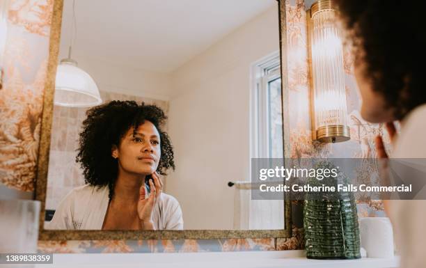 a beautiful woman examines her skin in a bathroom mirror - adult in mirror stock pictures, royalty-free photos & images