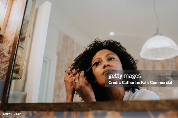 a woman applies mascara in a bathroom mirror - vestirse fotografías e imágenes de stock