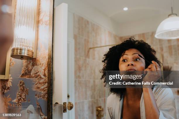 a woman applies mascara in a bathroom mirror - パウダールーム　女性 ストックフォトと画像