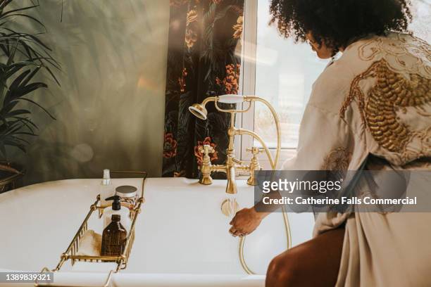 a woman checks the temperature of bath water by holding her hand under the faucet as the water runs - bubble bath foto e immagini stock