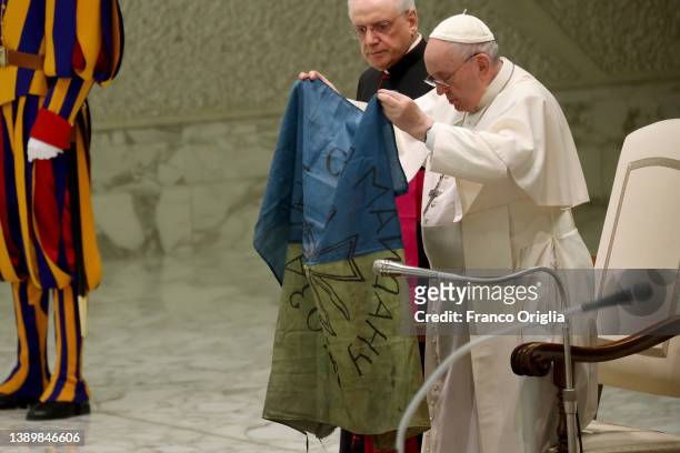 Pope Francis holds unfurled the faded and war-stained flag from that martyred city of Bucha and holds it up for all to see during his weekly general...