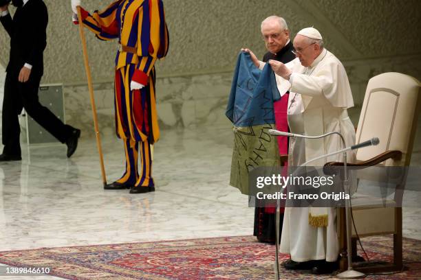 Pope Francis holds unfurled the faded and war-stained flag from that martyred city of Bucha and holds it up for all to see during his weekly general...