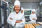 African American woman bakers looking at camera..Chef  baker in a chef dress and hat, cooking together in kitchen.She takes fresh baked cookies out of modern electric oven in kitchen.