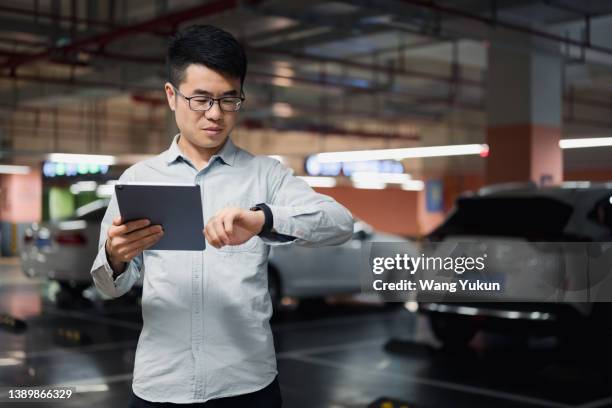 a male parking lot attendant is using an ipad while standing in an underground parking garage, he is looking at his smart watch - valet stock-fotos und bilder