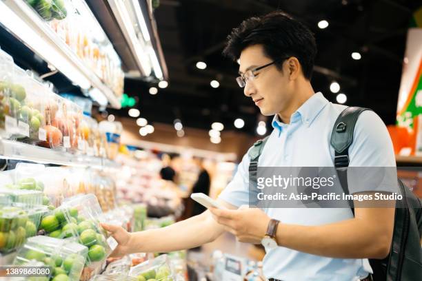 young asian man using smart phone  and choosing fresh organic fruits in supermarket. - people shanghai stockfoto's en -beelden