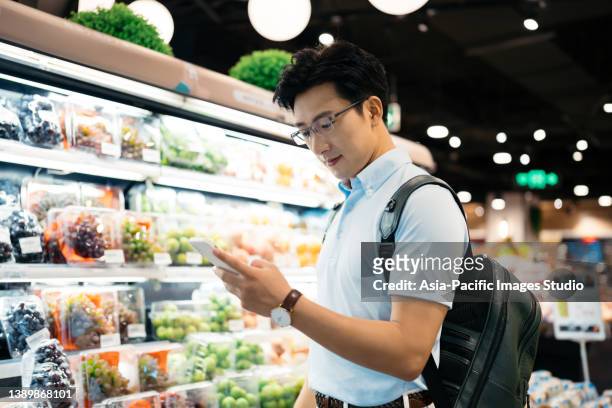 young asian man using smart phone  and choosing fresh organic fruits in supermarket. - people shanghai stockfoto's en -beelden