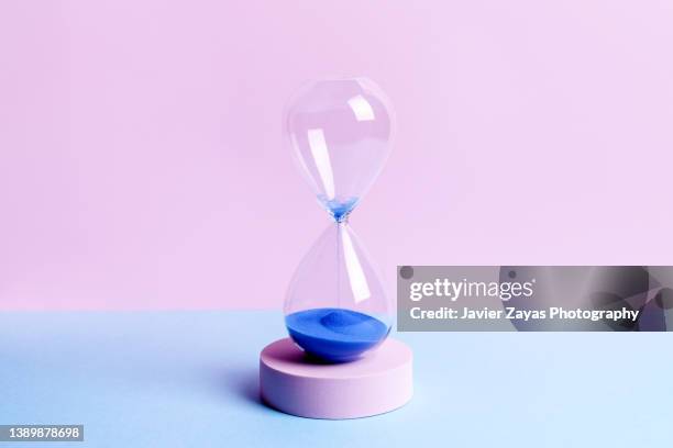 blue colored sand hourglass on blue and pink background - sand clock stock-fotos und bilder