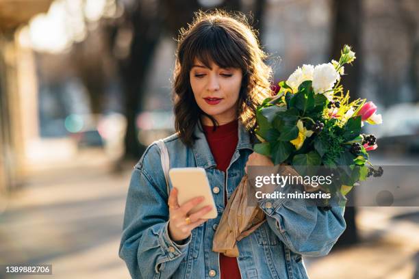 femme heureuse tenant des fleurs et utilisant un téléphone portable à l’extérieur - rencontre sur internet photos et images de collection