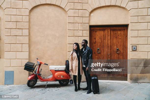 una pareja de jóvenes adultos está parada en una calle de siena cerca de un viejo scooter, esperando un taxi - women black and white motorcycle fotografías e imágenes de stock