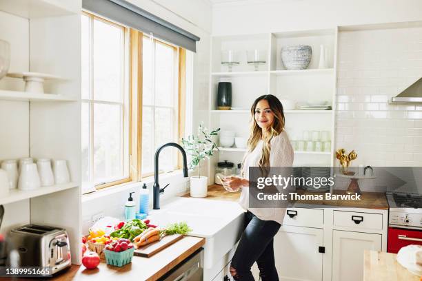 medium wide shot portrait of smiling woman standing by sink in kitchen - indian food bildbanksfoton och bilder