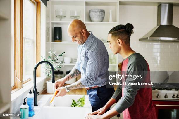 medium wide shot of smiling father and son washing fresh vegetables in kitchen sink - modern manhood stock pictures, royalty-free photos & images