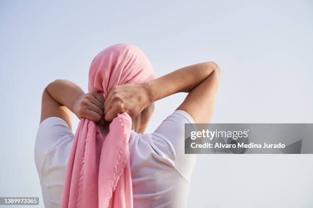 unrecognizable female cancer patient putting a pink scarf on her head with the sky in the background. concept of fighting and beating cancer. - borstkanker stockfoto's en -beelden