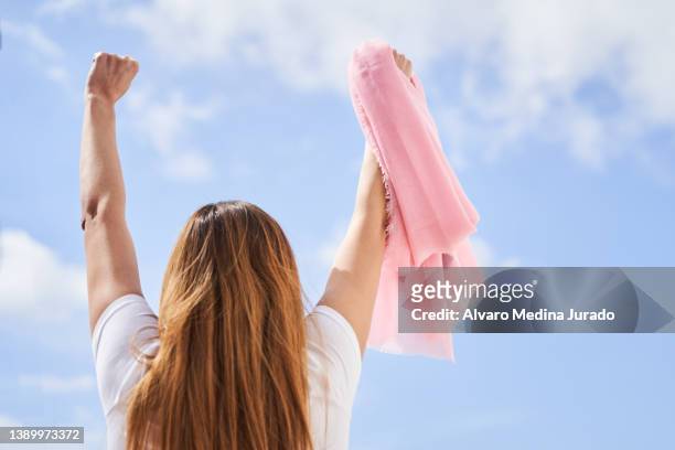 unrecognizable female cancer patient with pink handkerchief in hand and long hair, on her back with arms raised in victory sign with sky in background. concept of fighting and beating cancer. - best bosom fotografías e imágenes de stock