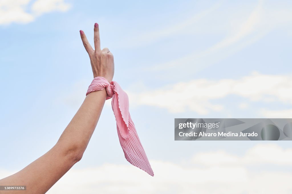 Close up shot of a female cancer patient's hand in victory sign with a pink scarf tied to her wrist. Concept of fighting and beating cancer.