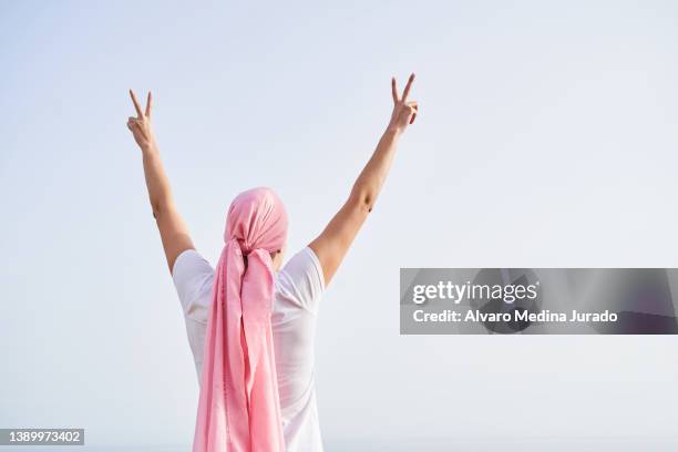 unrecognizable female cancer patient with pink headscarf on her head, back with arms raised in victory with sky in background. concept of fighting and beating cancer. - cancer research institute stock pictures, royalty-free photos & images