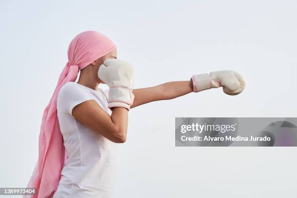 unrecognizable female cancer patient with pink headscarf, placed in punching position with boxing gloves in her hands as a sign of combat. concept of fighting and beating cancer. - best bosom fotografías e imágenes de stock