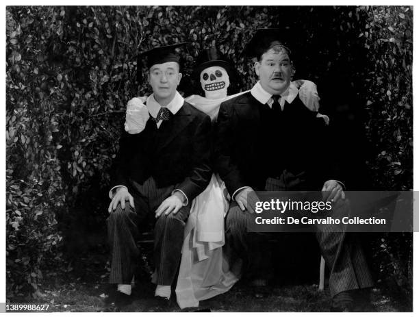 Actors Stan Laurel and Oliver Hardy sit on a park bench wearing their graduation gowns and beside a ghost hugging them from behind, in a scene from...