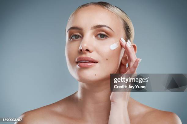 photo de studio d’une jolie jeune femme appliquant de la crème hydratante sur son visage sur un fond gris - applying stock photos et images de collection