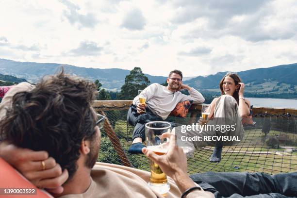 over the shoulder view of a young latino man lying on a net at a glamping site with his friends enjoying the scenery - beer glasses stock pictures, royalty-free photos & images