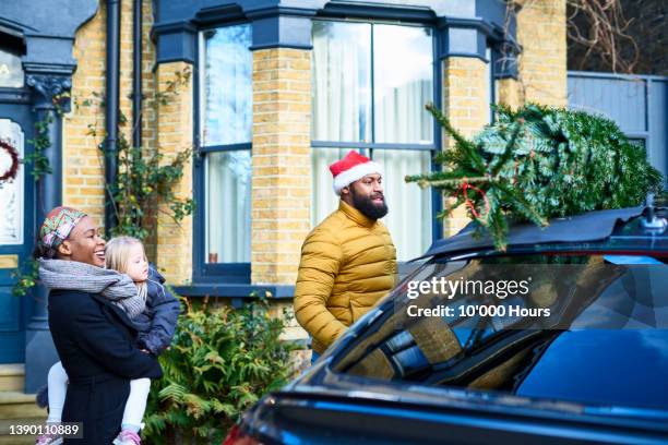 mid adult man in santa hat looking at christmas tree on roof of car outside house - sapin de noel humour photos et images de collection