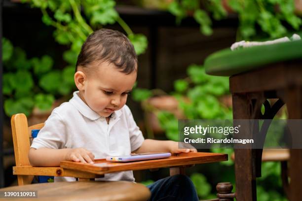 3-year-old latino boy is sitting in a highchair for a baby in a restaurant looking at his cell phone while waiting for his lunch - 2 year old child imagens e fotografias de stock